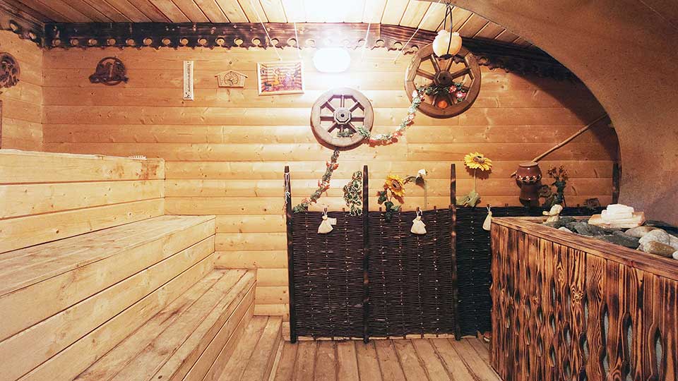 На фото: небольшое помещение сауны, справа - каменка, слева - полки, полы, стены, полки и потолок обшиты строганой доской, дальняя стена декоративно украшена, на стене - термометр