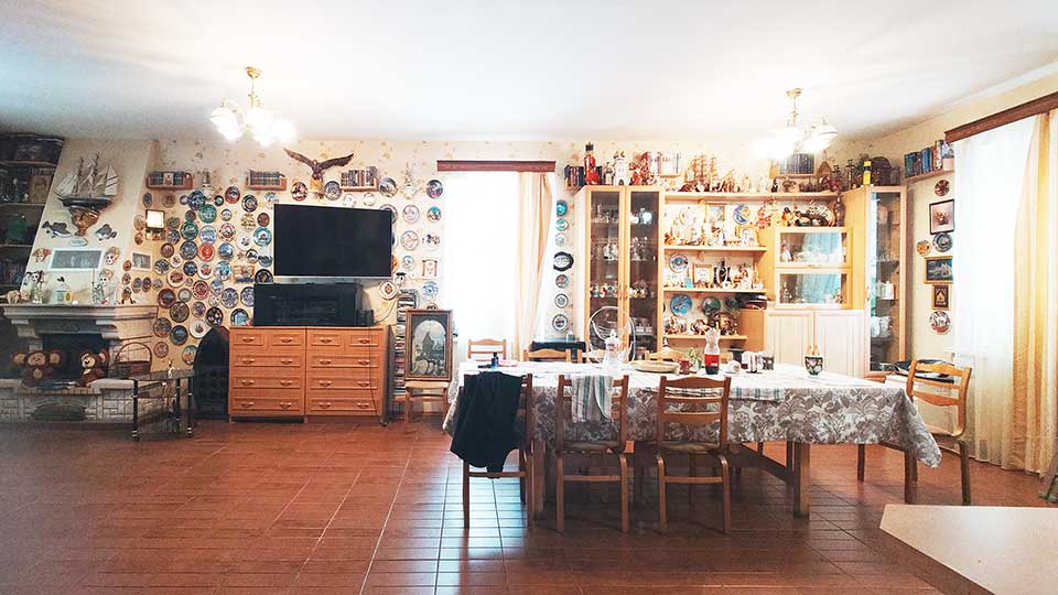 На фото: часть просторного светлого помещения - гостиной, полы - плитка, стены оклеены обоями, видно окно в дальней и правой стене, у дальней стены - сервант с посудой, небольшой комод, телевизионная панель на стене над комодом, действующий камин, все стены украшены декоративной посудой, по центру - обеденный стол со стульями вокруг него, стол накрыт скатертью, на столе - обеденная посуда, на потолке - две люстры