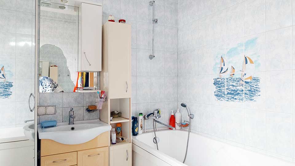 На фото: помещение ванной комнаты, стены - плитка на полную высоту, справа у стены - ванна со смесителем и душем, прямо у стены керамическая раковина на тумбе со своим смесителем, сверху, справа и слева - шкафчики, на верхнем шкафчике - зеркальная дверца