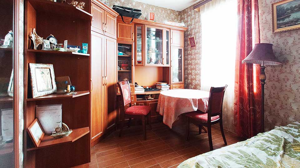 На фото: жилая комната с большим окном, полы - плитка, стены оклеены обоями, справа в углу - кровать, рядом с кроватью - торшер, по левой и дальней стене - мебельная стенка с одежным и посудным шкафом и открытыми полками,  у окна - круглый стол и два стула