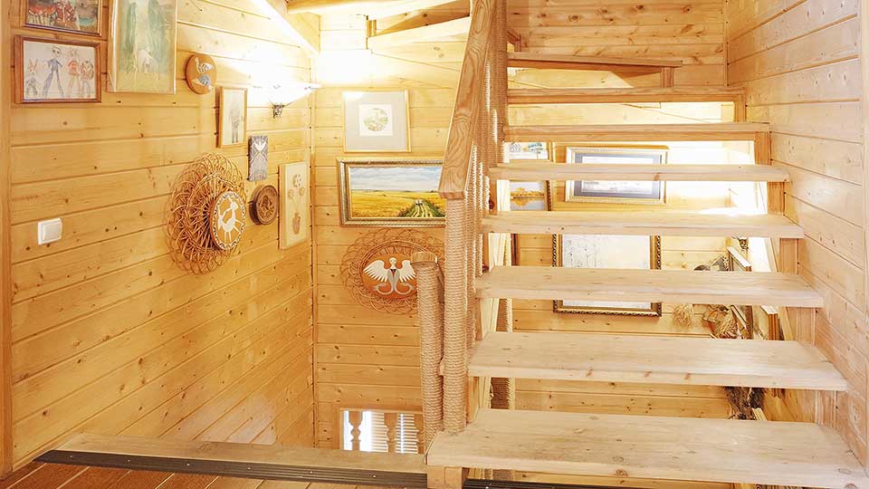 На фото: лестничный пролет деревянной двухпролетной лестницы, деревянные ступени, деревянные перила, стены пролета облицованы строганой доской, установлены настенные светильники, на стенах - декоративные изделия