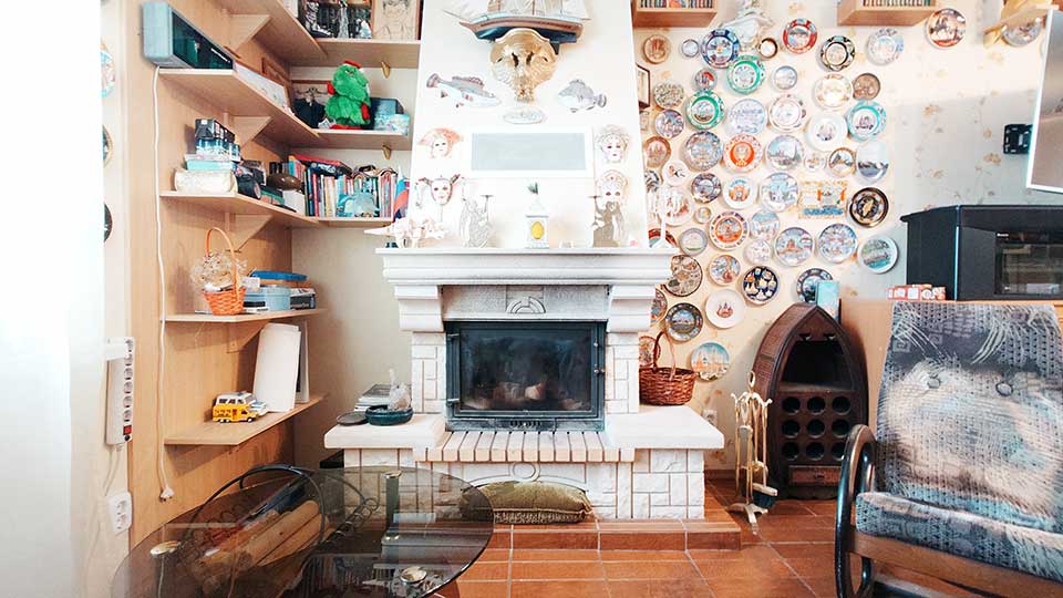 На фото: пространство у камина, камин в центре фото, обложен камнем, зона горения закрыта жаропрочным стеклом, слева - окно, у окна на стене полки с житейской мелочью, справа от камина на стене панно из декоративных тарелок, справ от камина на полу - кресло-качалка