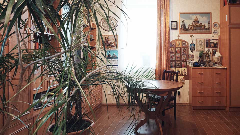 На фото: часть помещения жилой комнаты, полы - плитка, стены оклеены обоями, слева от окна по левой стене - мебельная стенка из различных шкафов и полок, справа от окна комод и платяной шкаф, перед окном - круглый стол со стулом, слева у шкафа - пальма в кадке