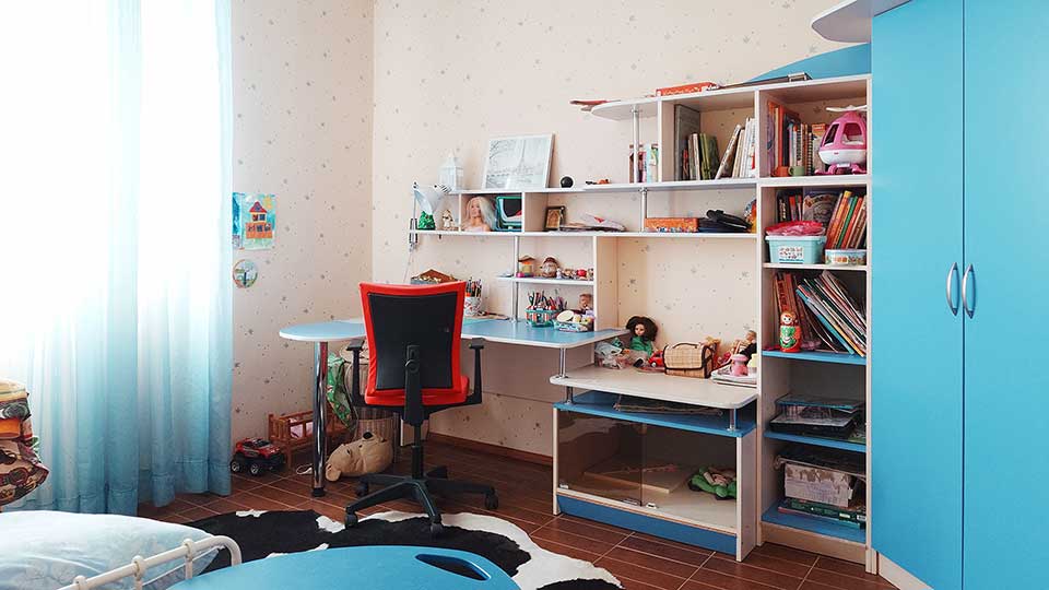 На фото: часть помещения жилой комнаты - детской, полы - плитка, стены оклеены обоями, слева от окна у стены - кровать, вдоль правой стены - детская мебельная стенка в голубых тонах, прямо в углу - письменный стол и офисное кресло на колесиках