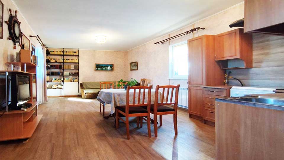 На фото: просторная жилая комната (вид от входа в комнату), полы - ламинат, стены оклеены обоями, потолок окрашен, справа - оборудованная полноценная кухня с мойкой, плитой, вытяжкой, тумбами, навесными шкафами и шкафом-пеналом, посредине комнаты - обеденный стол и четыре стула вокруг него, слева у стены - туба с телевизором и полками,  у дальней стены - диван и стеллаж с книгами, два окна дают много света с двух сторон