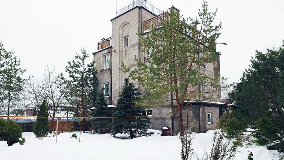 На фото: фасад трехэтажного жилого дома, стены оштукатурены, плоская эксплуатируемая кровля, перед домом - земельный участок с газоном и деревьями, по земельному участку к дому проходит газовая труба по металлическим столбам, на дальнем плане - забор из металлопрофиля по металлическим столбам, за забором - дом на соседнем участке.